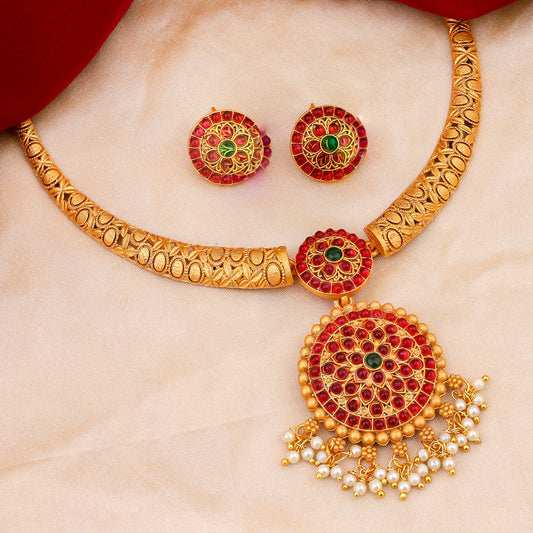 A gold necklace with intricate designs and red and green gemstones, accompanied by matching earrings, displayed on a beige fabric background.