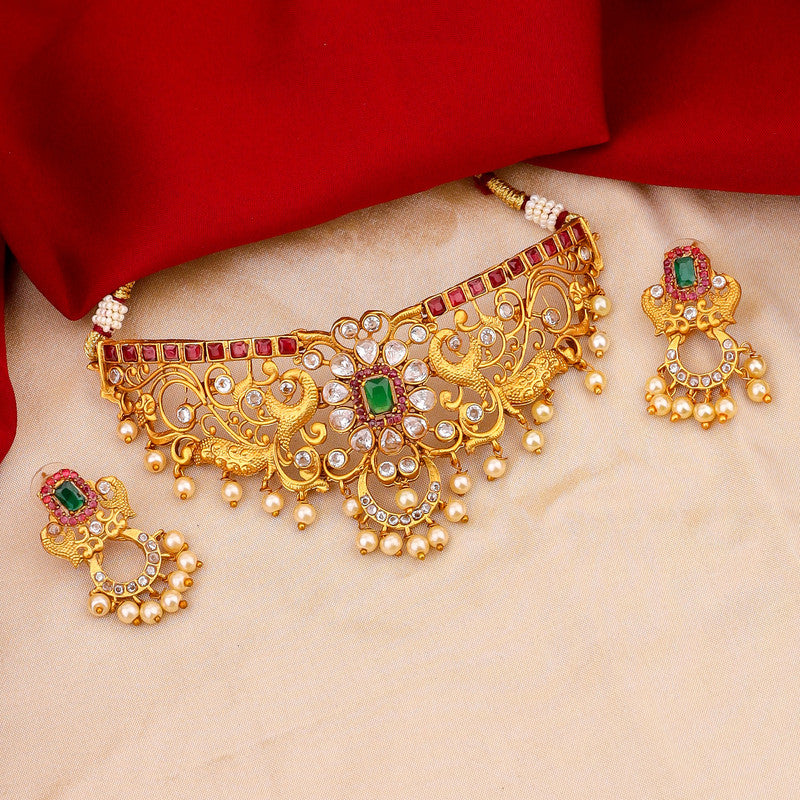 A set of ornate gold jewelry with red and green gemstones, featuring a necklace and matching earrings, displayed on a beige surface with a red fabric background.