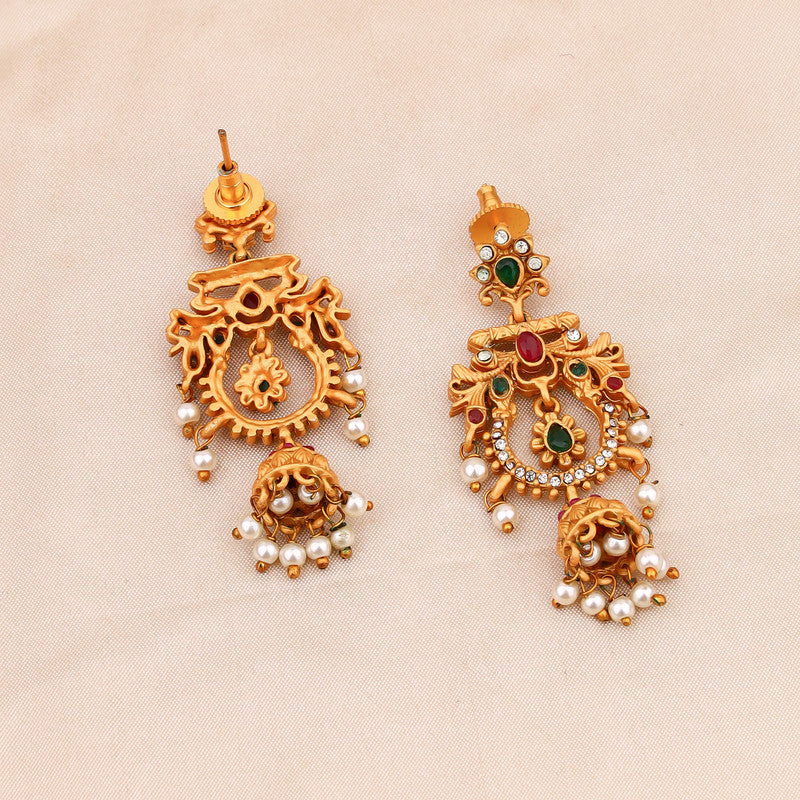 A pair of ornate gold earrings with intricate designs, featuring red and green gemstones and pearl dangles, displayed on a beige fabric background.