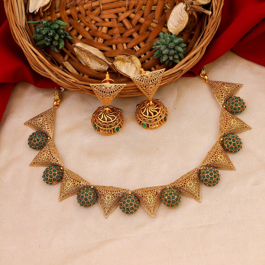 A set of intricate gold jewelry with green accents, including a necklace with triangular and circular designs, and matching earrings, displayed on a beige fabric with a wicker basket and red cloth in the background.