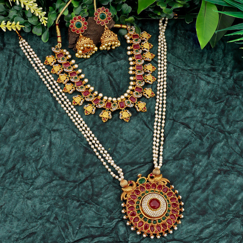 A set of traditional Indian jewelry on a green textured background, featuring a long necklace with a large circular pendant, a shorter necklace with intricate designs, and a pair of matching earrings. The jewelry is adorned with red and green gemstones and gold detailing, surrounded by green leaves.