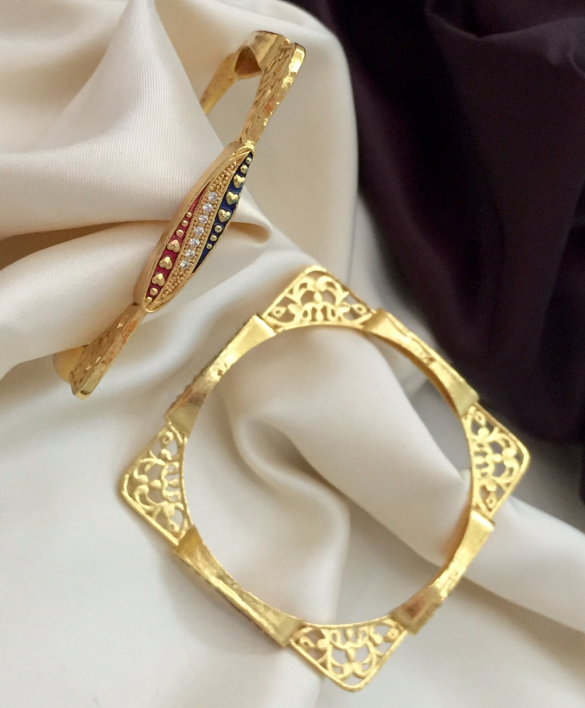 Two gold bangles, one with intricate cut-out designs and the other adorned with multicolored gemstones.