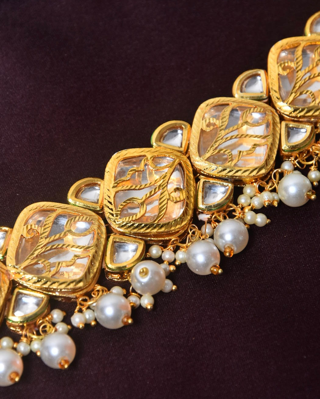 A gold choker necklace with intricate designs, featuring square-shaped elements and adorned with white pearls and small reflective stones.