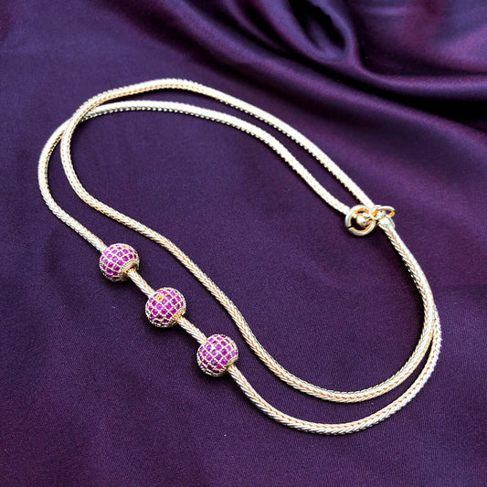 A gold necklace with three spherical pink gemstone-studded beads, displayed on a dark purple fabric background.