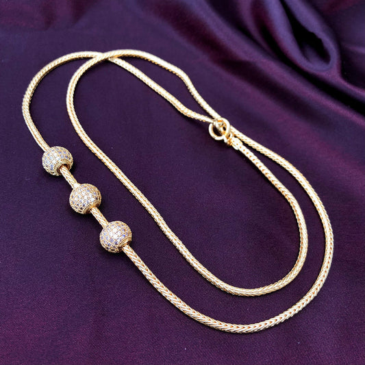 A gold necklace with three spherical, diamond-encrusted beads displayed on a dark purple fabric background.