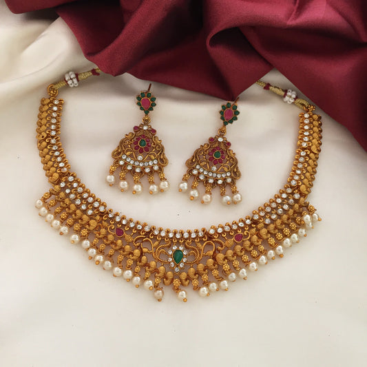 A traditional gold necklace set with intricate designs, adorned with pearls and red and green gemstones, accompanied by matching earrings, displayed on a white and maroon fabric background.