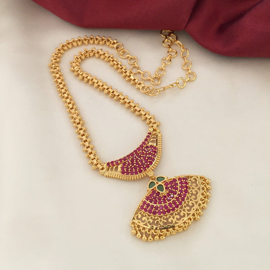 A gold necklace with intricate designs, featuring two crescent-shaped pendants adorned with red and green gemstones, displayed on a light-colored surface with a red fabric in the background.