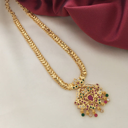 A gold necklace with a detailed pendant featuring red, green, and white gemstones, displayed on a light surface with a red fabric background.