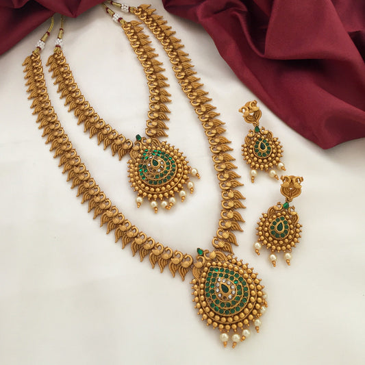 A set of traditional gold jewelry consisting of two necklaces and a pair of earrings. The necklaces feature intricate designs with green and white stones and pearl accents, with a large pendant in the center. The earrings match the necklaces with similar designs and dangling pearls. The jewelry is displayed on a white surface with a maroon fabric in the background.