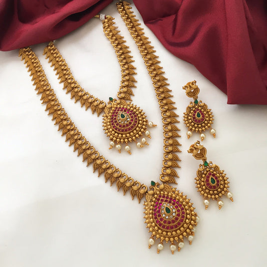 A set of traditional gold jewelry consisting of two necklaces and a pair of earrings, adorned with red and green gemstones and pearl accents, displayed on a white surface with a red fabric backdrop.