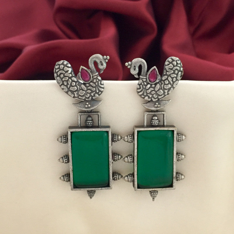 Silver earrings with peacock designs at the top and rectangular green stones at the bottom, displayed against a white and red fabric background.