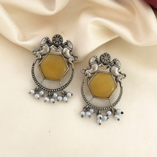 A pair of silver earrings featuring a hexagonal yellow stone, two horse figures, and dangling white pearls.